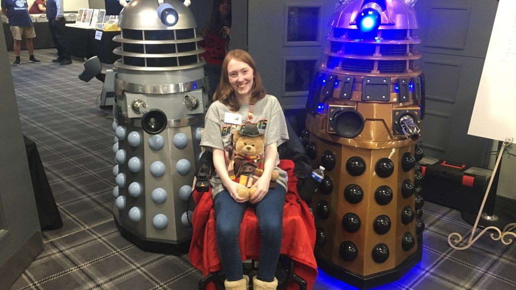 Erin, a white woman with ginger hair, sits in a powerchair. She is smiling. She is holding a Build-a-Bear dressed as the Fourth Doctor on her lap. There are two life-sized Dalek replicas either side of her.