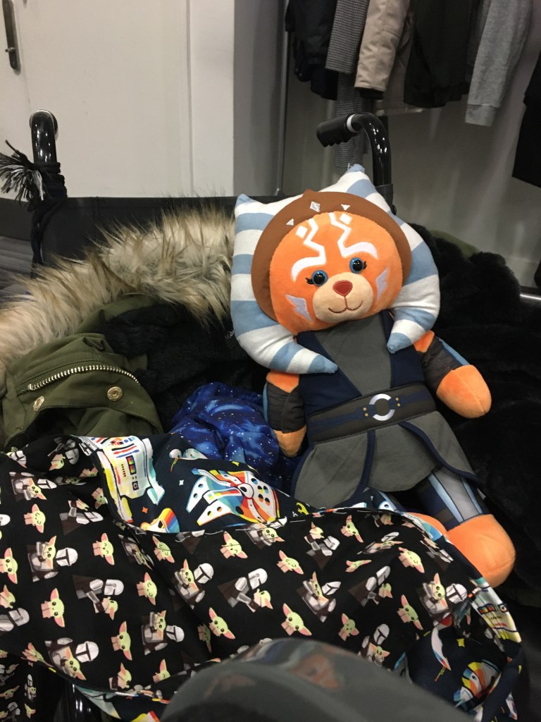 A black, Red Cross transit wheelchair. On the wheelchair is a coat, a Star Wars-printed bag, and a Build-a-Bear plush of Ahsoka Tano.